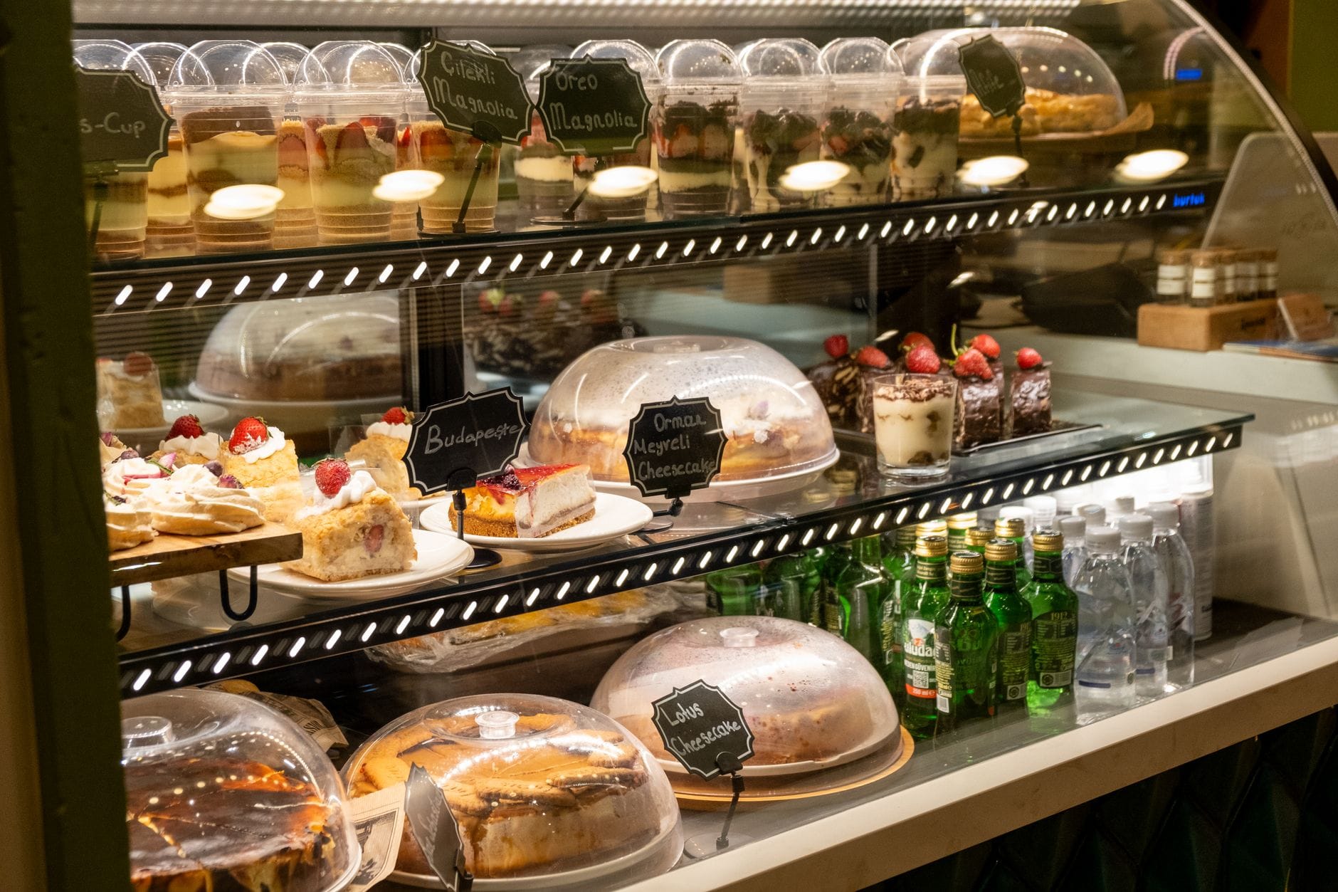 Glasvitrine gefüllt mit Kuchen und Gebäck, beschrifteten Desserts und Flaschengetränken in einem gemütlichen, einladenden Café in Budapest.