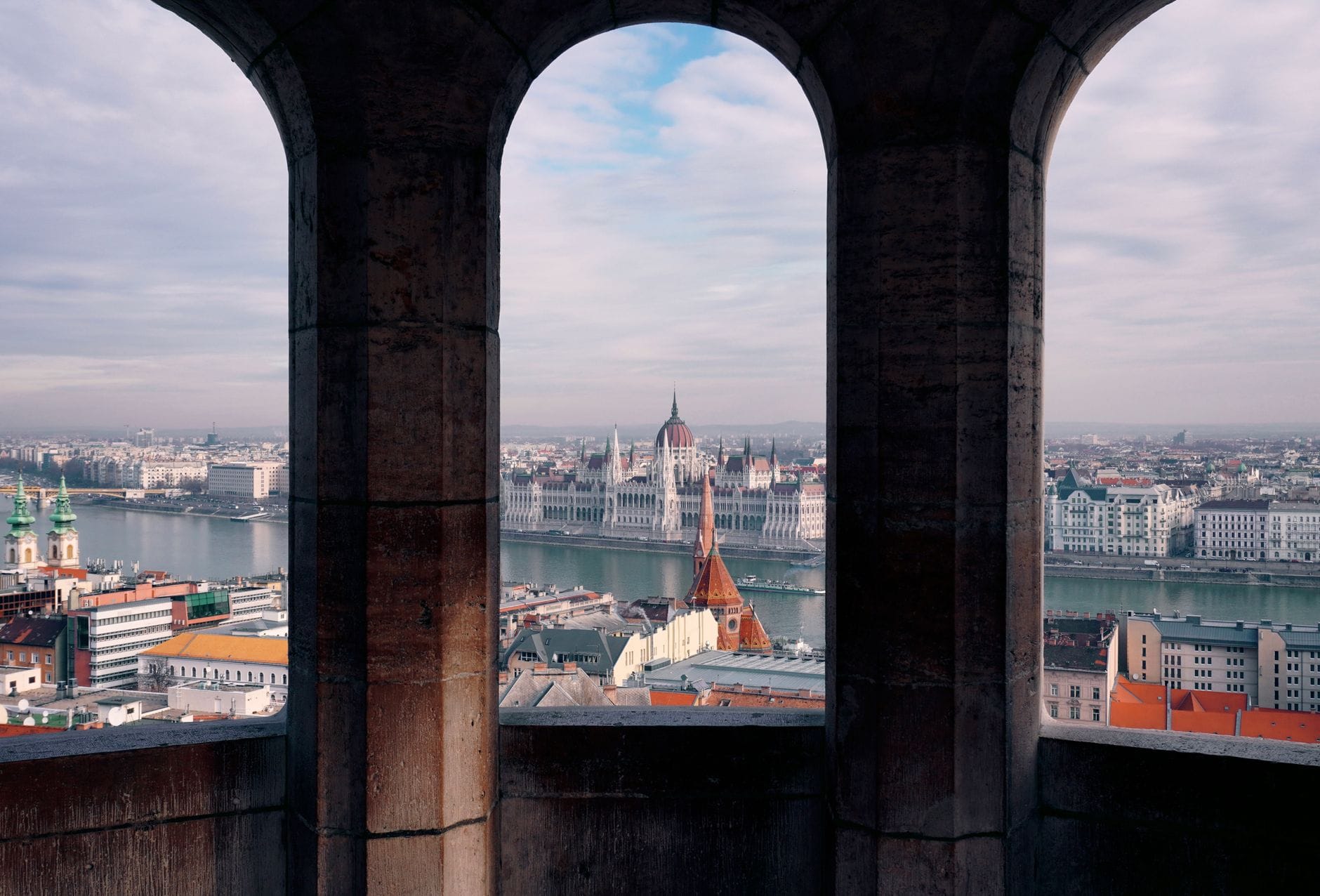 Blick durch gewölbte Steinfenster auf die Donau und das Ungarische Parlament in Budapest an einem hellen Tag.