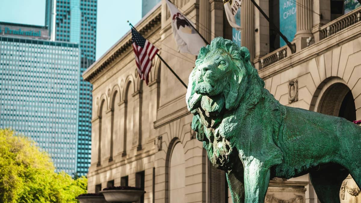 Icónica estatua de león de bronce frente al Art Institute of Chicago con banderas, edificio histórico y rascacielos de la ciudad visibles al fondo.