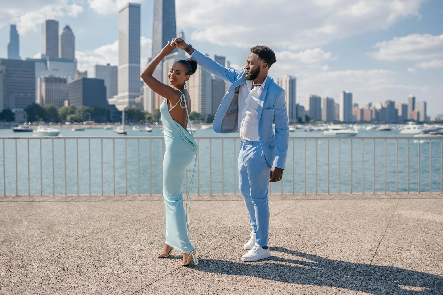 Um casal estiloso dança num calçadão à beira-mar com o skyline de Chicago e barcos ao fundo em um dia ensolarado.