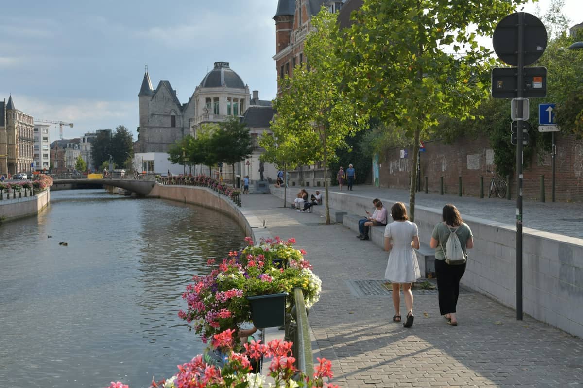 Canal in Ghent lined with blooming flowers, historic stone buildings, a walking path, and people enjoying the summer afternoon.