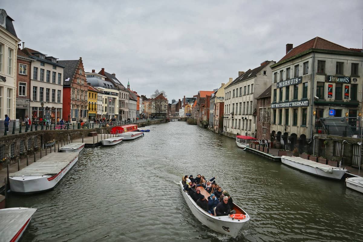 Open canal boat tour with passengers moves through Ghent's canal lined with colorful medieval buildings on both sides under cloudy sky.