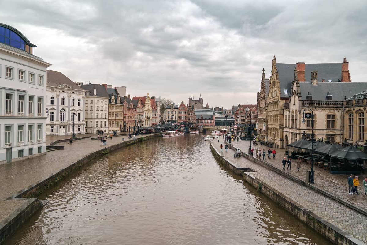 A wide view of Ghent's historic canal and medieval buildings under cloudy skies, with people walking along the riverside paths.