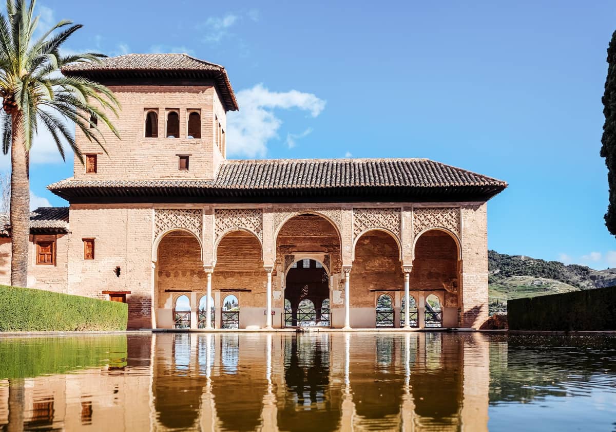 พระราชวัง Partal แห่ง Alhambra พร้อมต้นปาล์มและระเบียงโค้งที่สะท้อนในสระน้ำสี่เหลี่ยมผืนผ้ายาวใต้ท้องฟ้าสีฟ้าสดใส