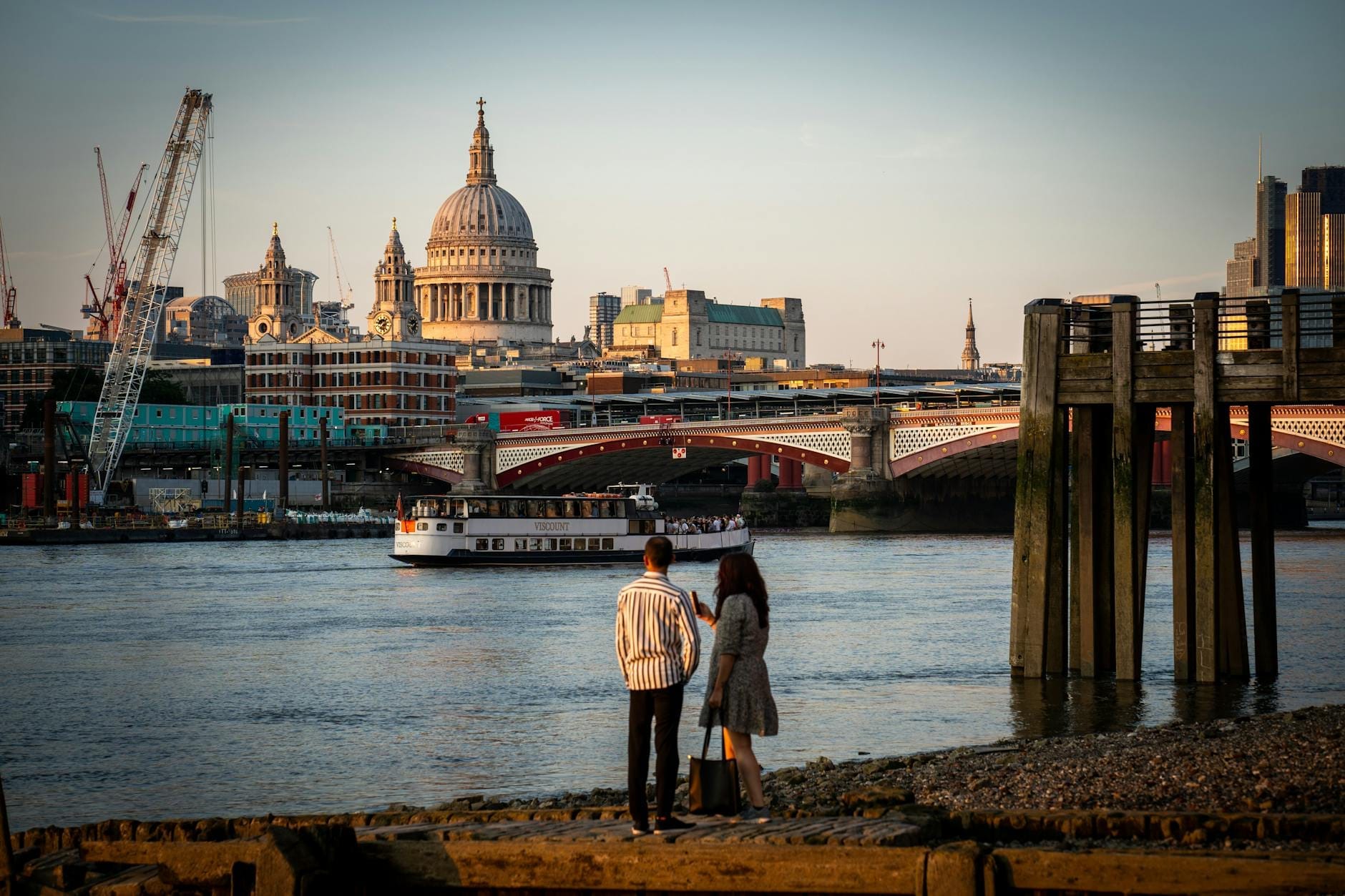 Um casal está à beira do Tâmisa ao pôr do sol, olhando em direção à Catedral de São Paulo e a um barco passando, com o skyline de Londres ao fundo.