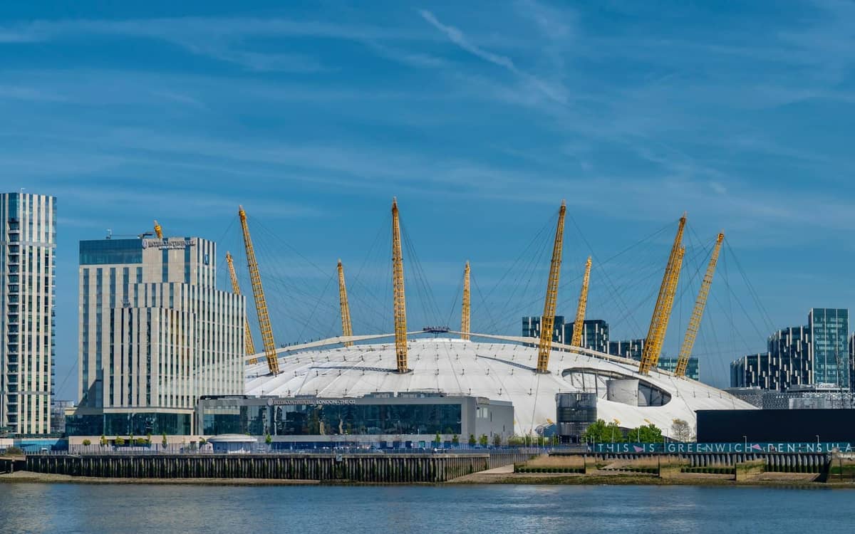 Vista diurna do O2 Arena em Londres com edifícios modernos e o rio Tâmisa em primeiro plano.