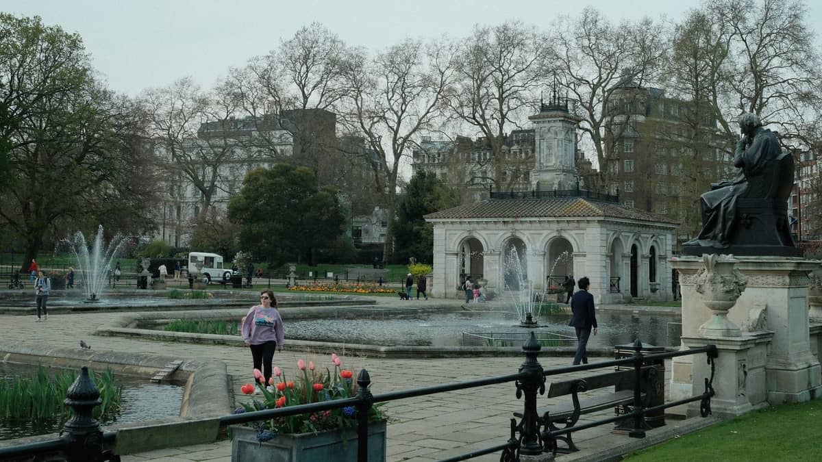 Pessoas passeiam pelos Italian Gardens em Hyde Park, com uma histórica casa de bombas, fontes, estátuas, tulipas e a arquitetura de Londres ao fundo.