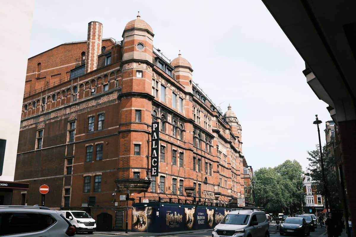 Vista da rua do histórico Palace Theatre em Londres com fachada de tijolos vermelhos, letreiros visíveis e carros estacionados do lado de fora durante o dia.