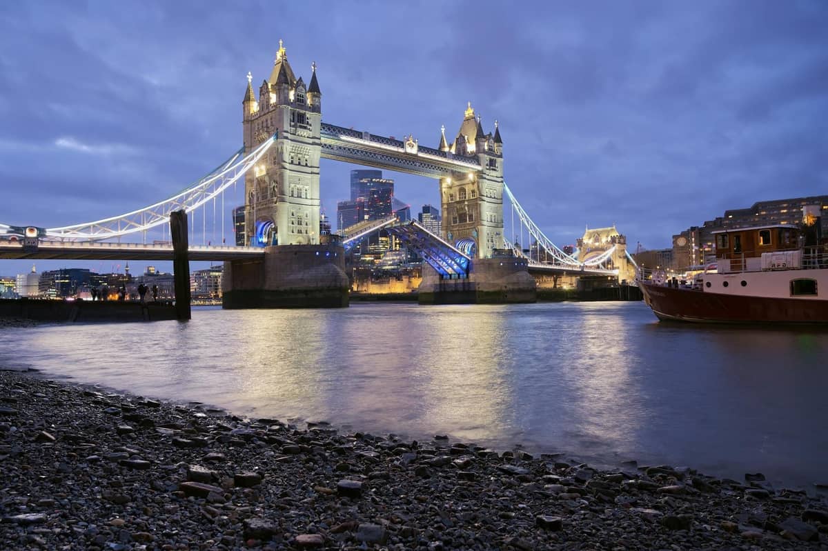 Tower Bridge iluminada ao anoitecer com as luzes da cidade refletidas no Rio Tâmisa e um barco próximo, capturando uma noite romântica em Londres.