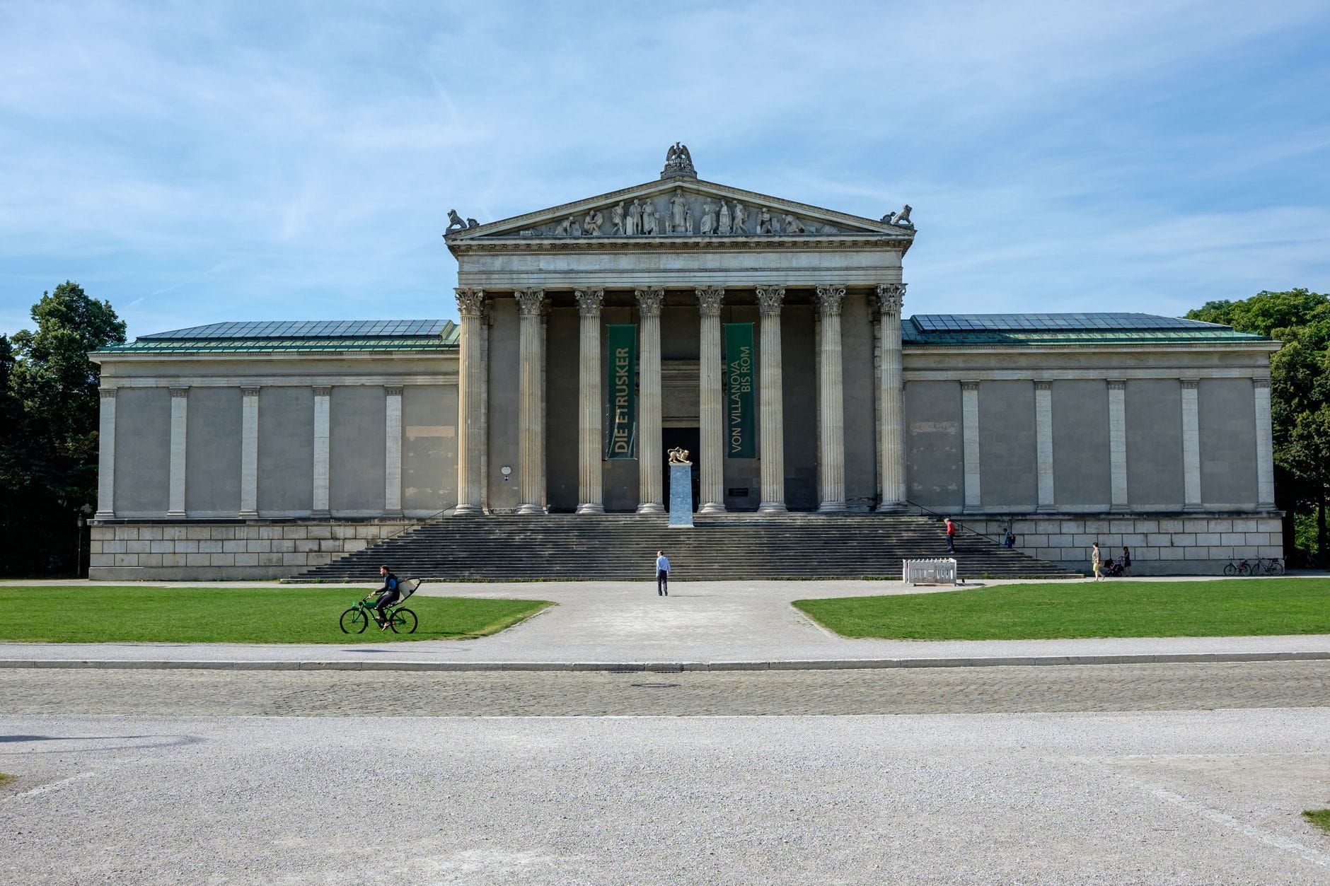 ด้านนอกสไตล์นีโอคลาสสิกของพิพิธภัณฑ์ Glyptothek ในมิวนิกในวันที่มีแดดสดใส พร้อมผู้คนเดินและปั่นจักรยานอยู่ด้านหน้า