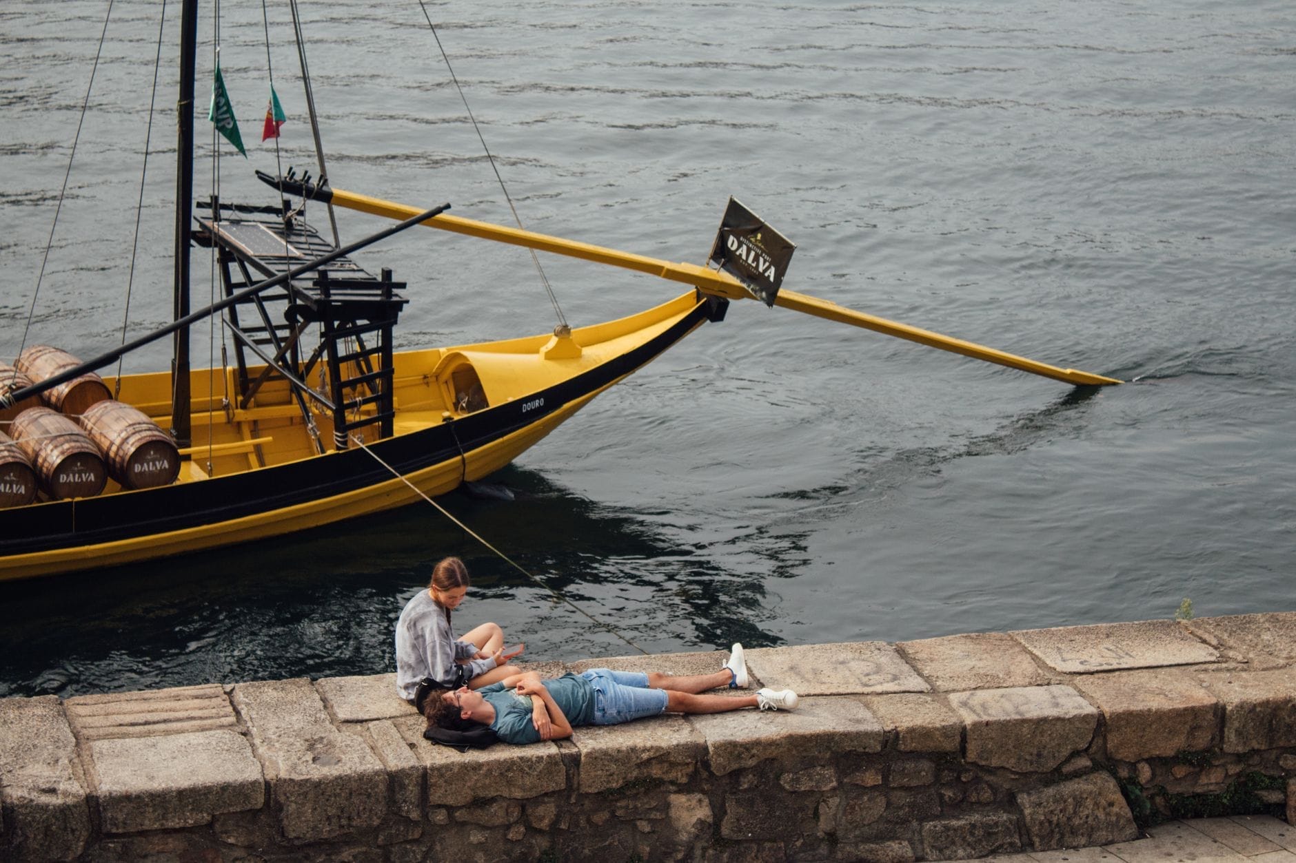 Um casal relaxando às margens do Rio Douro com um barco rabelo tradicional ao fundo, evocando romance e o charme único da cidade.