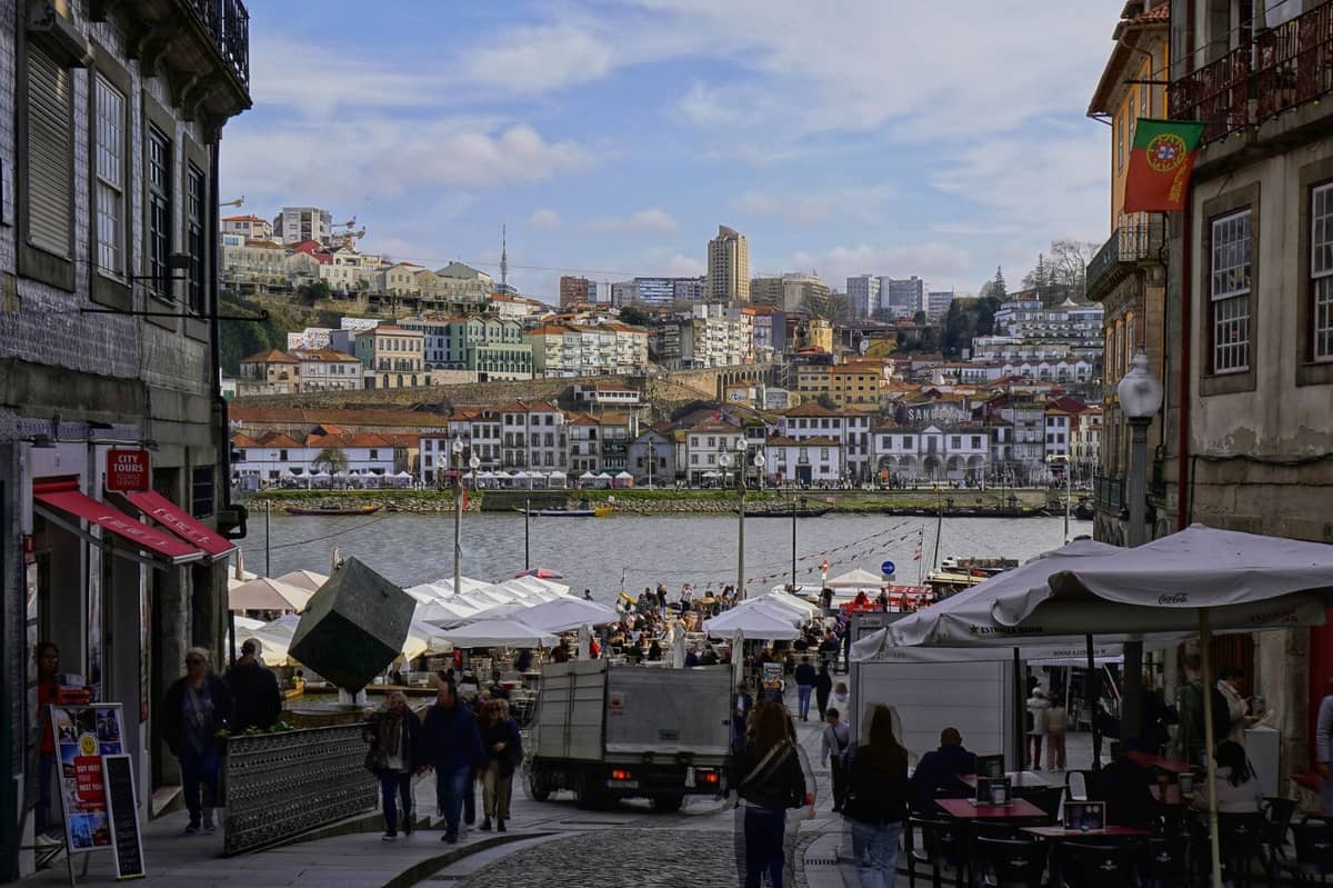 Vista de una calle que conduce al frente del río Douro en Porto con edificios coloridos, cafés al aire libre y personas paseando, capturando el ambiente animado e íntimo de la ciudad.
