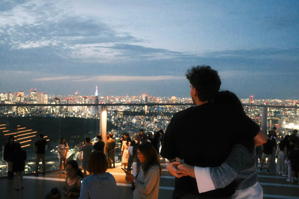 Couples and visitors enjoy sunset panoramic city views from a Tokyo observation deck, with city lights beginning to glow and a romantic atmosphere.