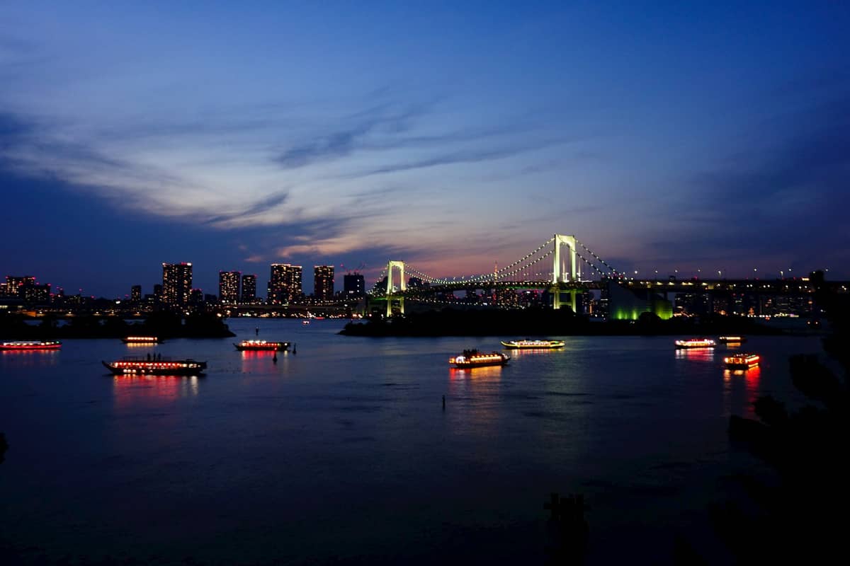 Multiple illuminated dinner cruise boats sail on Tokyo Bay at dusk with Rainbow Bridge and Odaiba skyline lit up in the background.