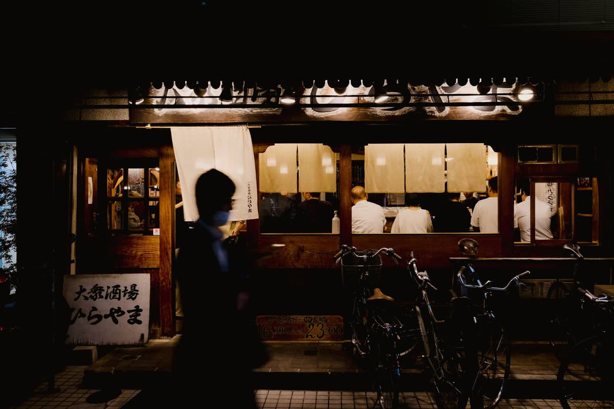 Nighttime view of a cozy Japanese restaurant with chefs behind a counter and warm lighting, seen from outside in Tokyo.