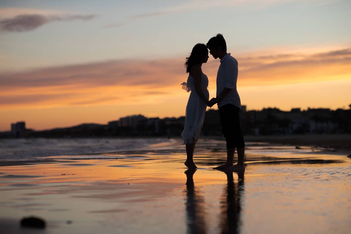 Couple se tenant la main et se penchant pour s'embrasser sur une plage de sable au coucher du soleil avec le paysage urbain en arrière-plan.