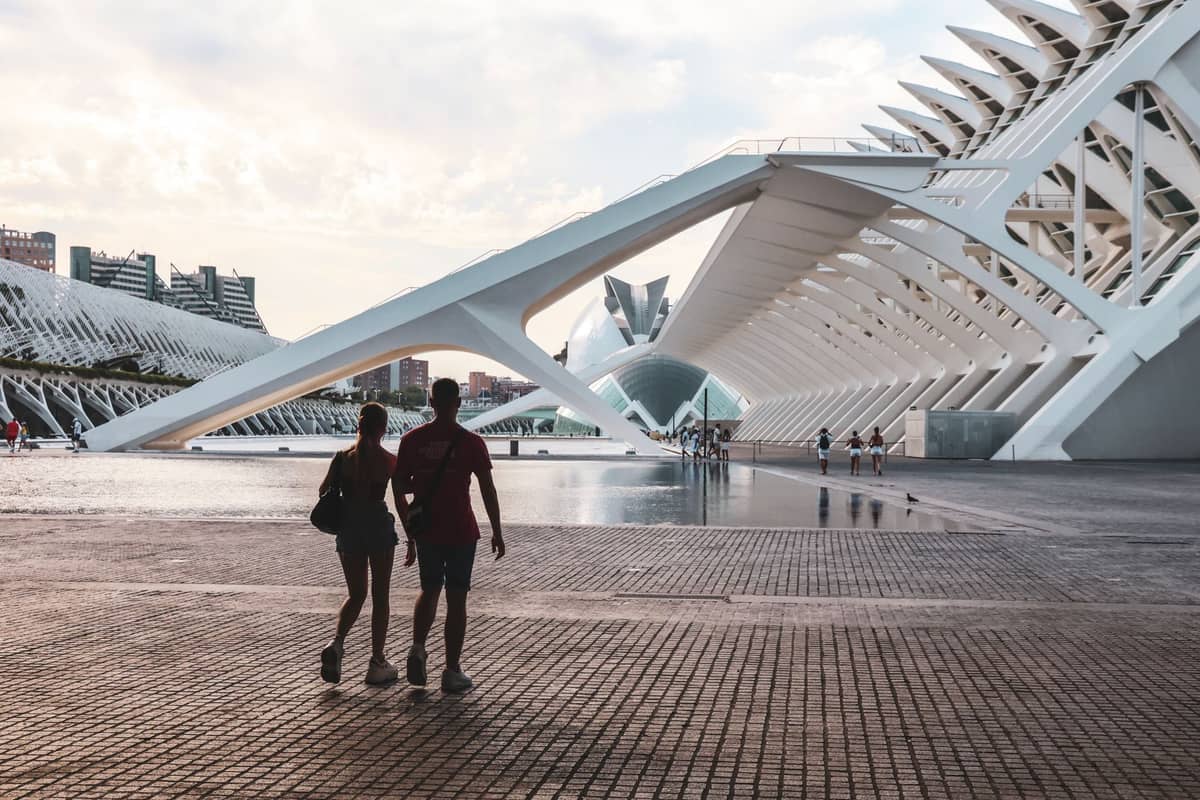 Una coppia passeggia vicino agli imponenti archi bianchi e agli edifici futuristici della Ciudad de las Artes y las Ciencias di Valencia sotto un soffice cielo serale.