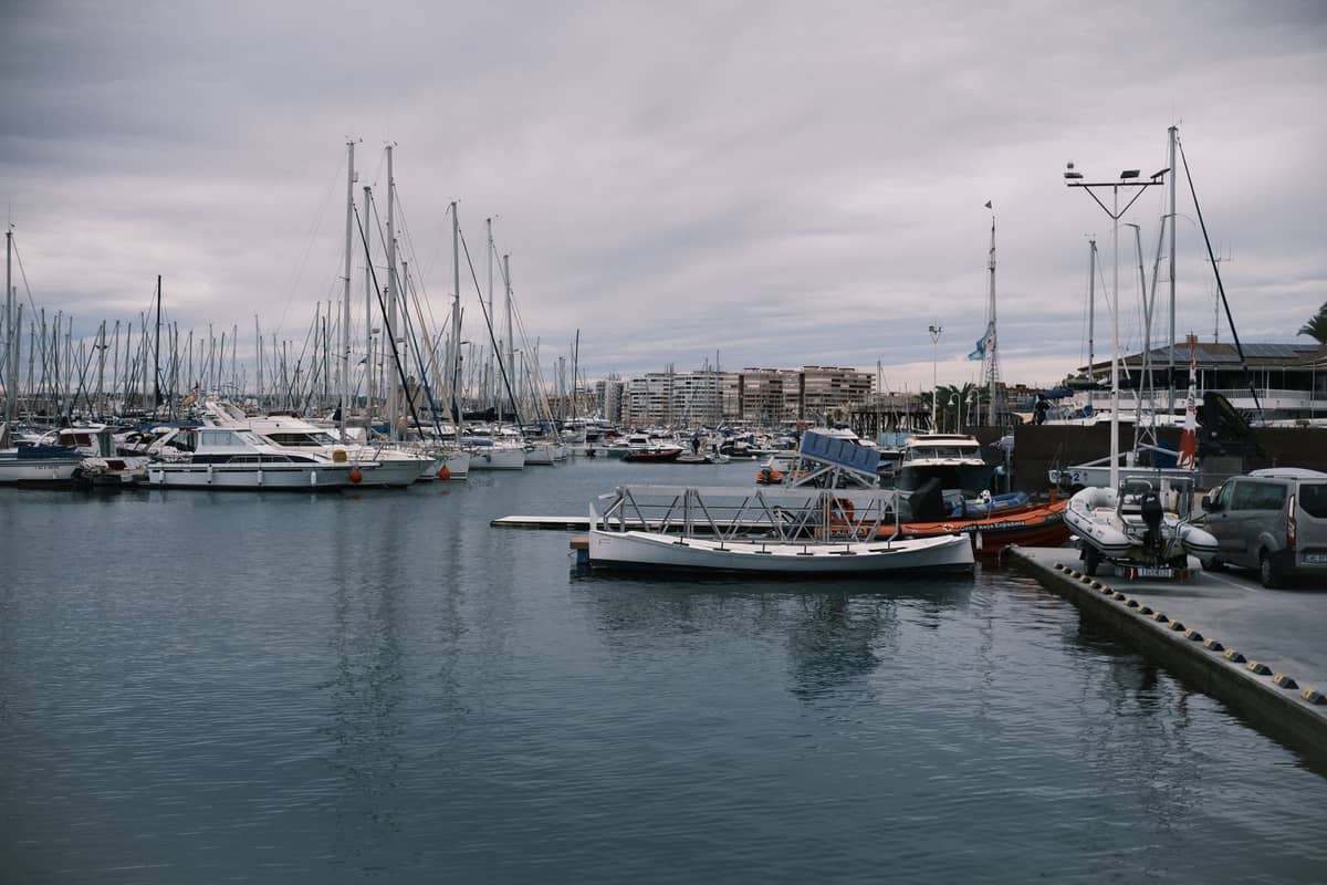 Vue du port de plaisance de Valencia avec de nombreux voiliers amarrés, l'eau, les quais et des bâtiments distants sous un ciel nuageux.