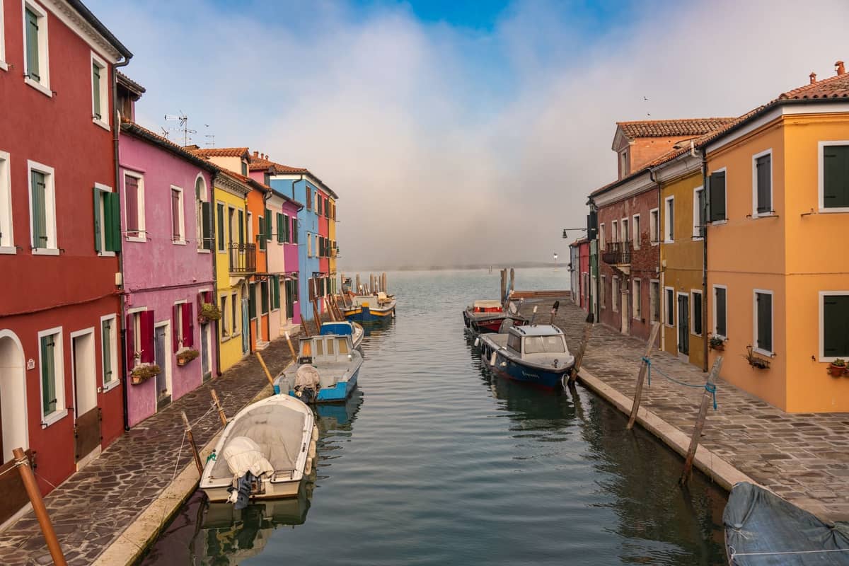 Kanal gesäumt von lebhaft bemalten Häusern in Ocker, Rosa, Blau und Rot mit kleinen Booten, die entlang des Wassers vertäut sind, in Burano, Venedig.
