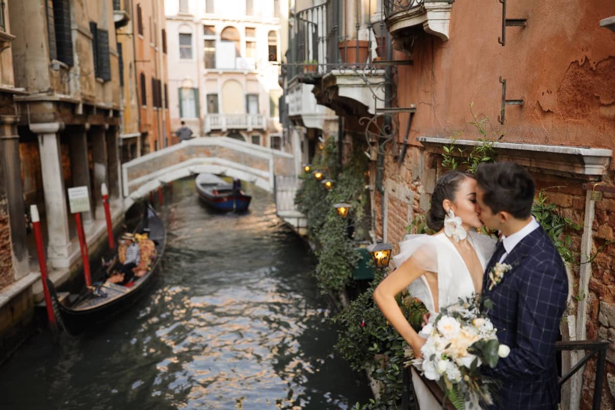 Ein Paar küsst sich an einem Venezianischen Kanal, im Hintergrund Gondeln und eine romantische Brücke, umgeben von historischen Gebäuden.