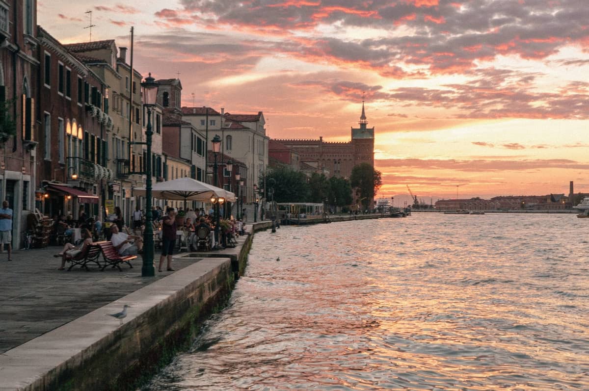 Venezianische Uferpromenade bei Sonnenuntergang mit Menschen, die vor Cafés sitzen und spazieren gehen, historische Gebäude entlang des Kanals und ein sanft rosa-orangefarbener Himmel.