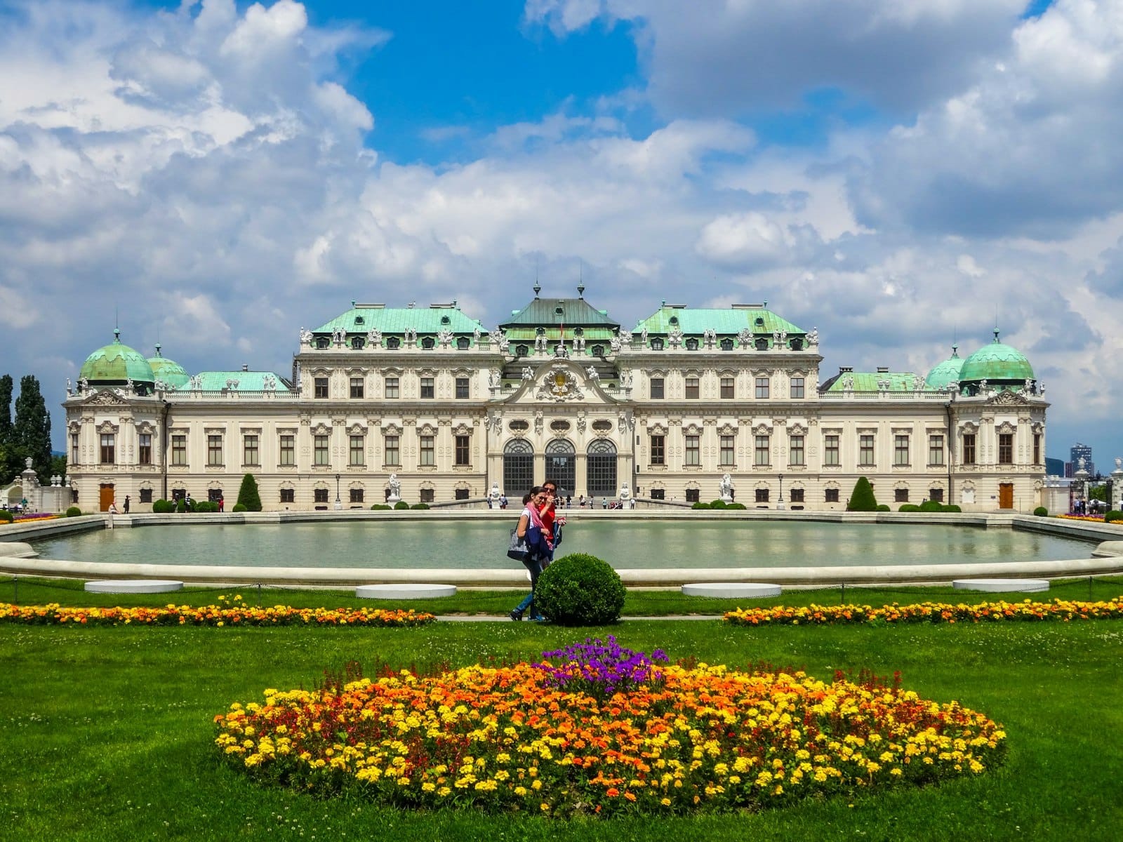 Ein Paar steht vor dem eleganten Belvedere-Palast in Wien, umgeben von lebhaften Gärten und einem Spiegelbecken an einem sonnigen Tag.