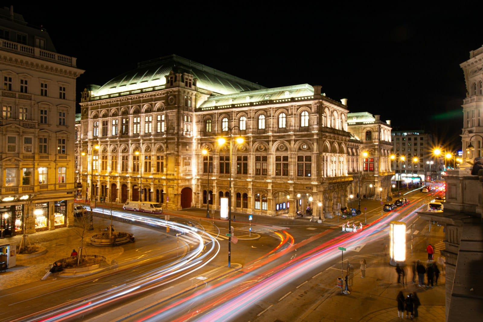 Nachtansicht der Wiener Staatsoper, warm beleuchtet mit belebter Straße, Lichtspuren und vorbeigehenden Menschen im Vordergrund.