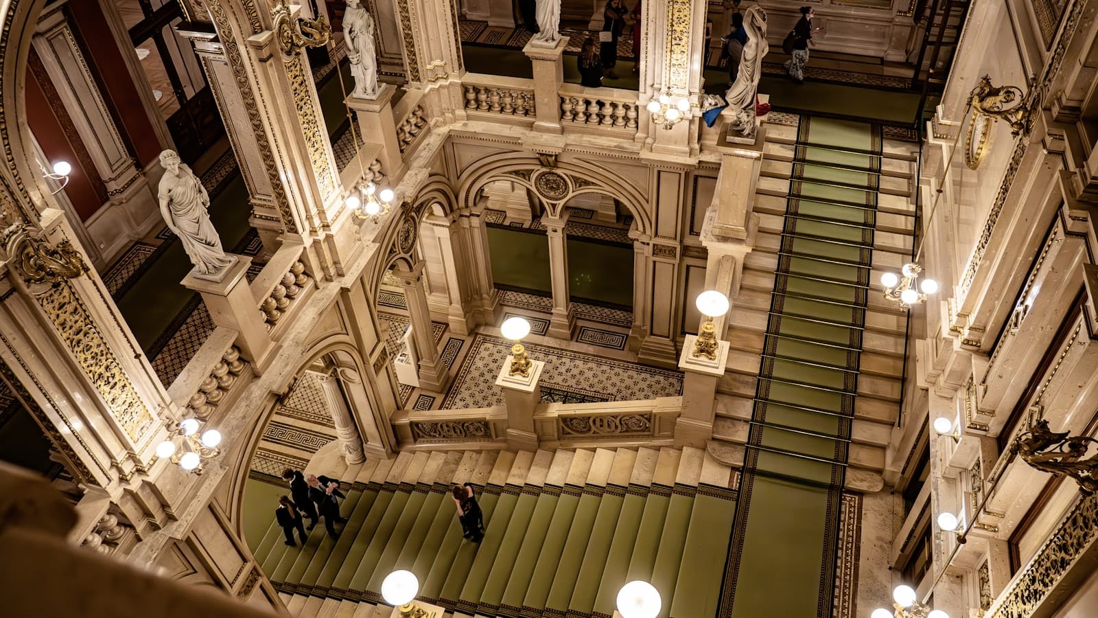 Prachtvolle große Treppe mit Statuen, dekorativen Leuchten und architektonischen Details im Inneren eines historischen Gebäudes in Wien, von oben betrachtet.