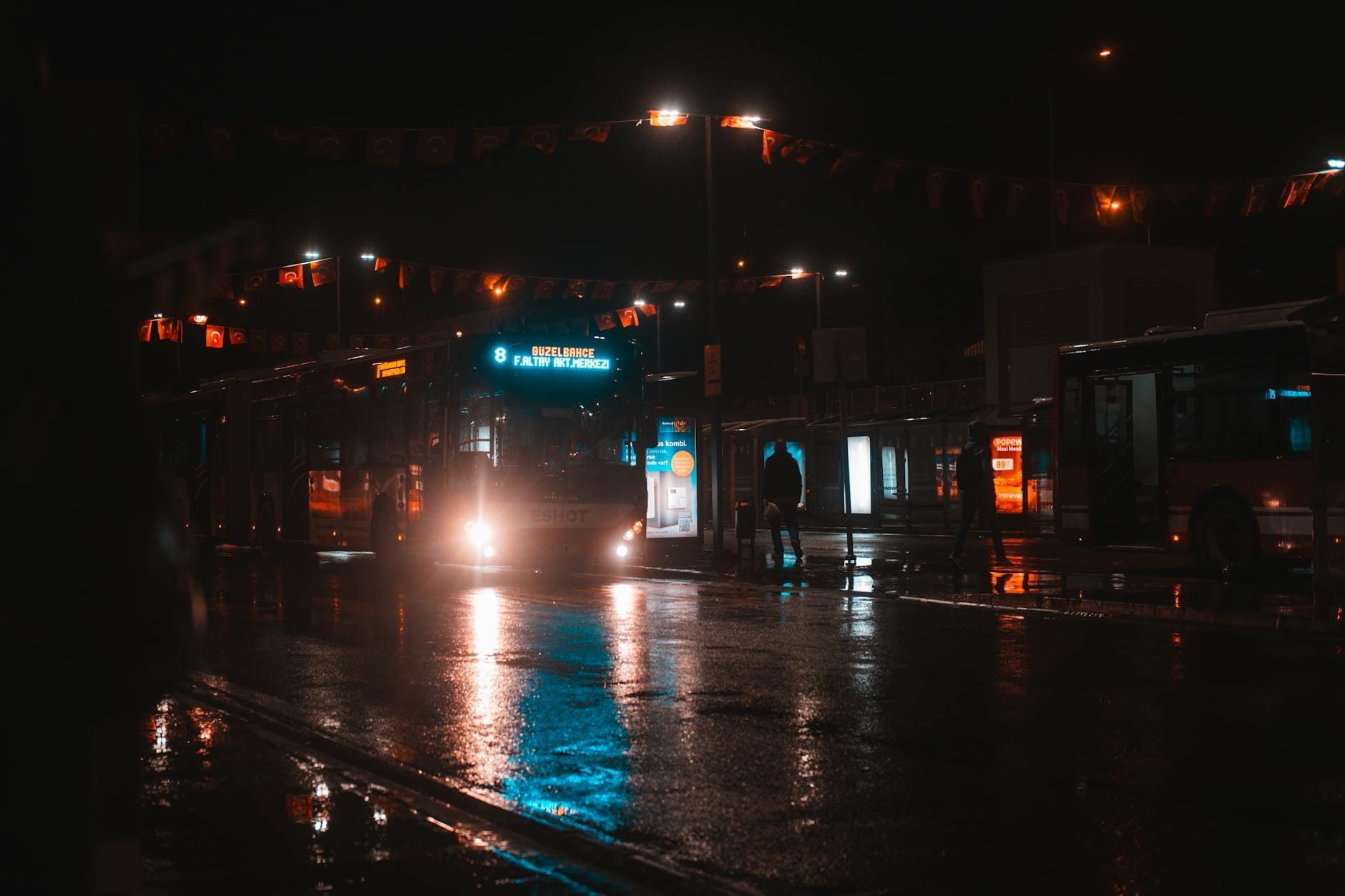 Night scene at a city bus terminal in the rain, with illuminated buses and wet pavement reflecting streetlights.