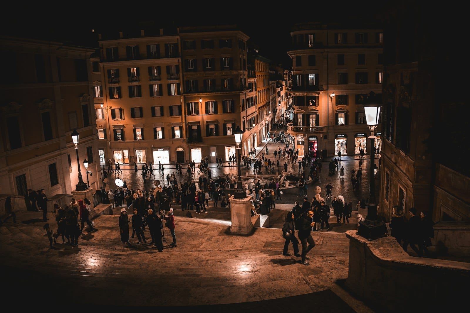 View from the Spanish Steps at night with people walking and gathering, and warmly lit historic buildings in Rome.