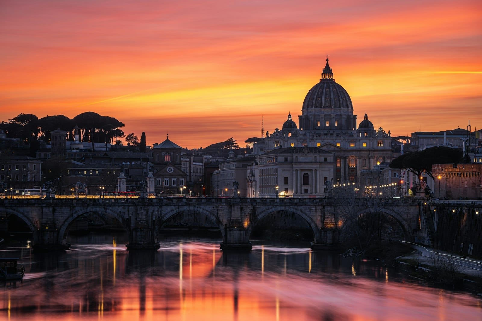 St. Peter's Basilica and the Tiber River in Rome glowing at sunset, with lights reflecting in the water and a dramatic, colorful sky.