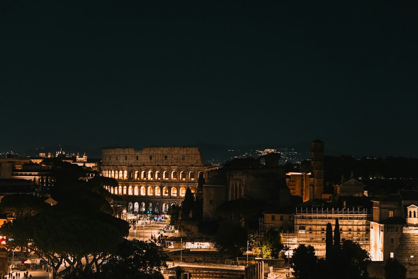A wide, beautifully lit night view of the Colosseum and surrounding Roman streets, with city lights and ancient ruins glowing under the dark sky.