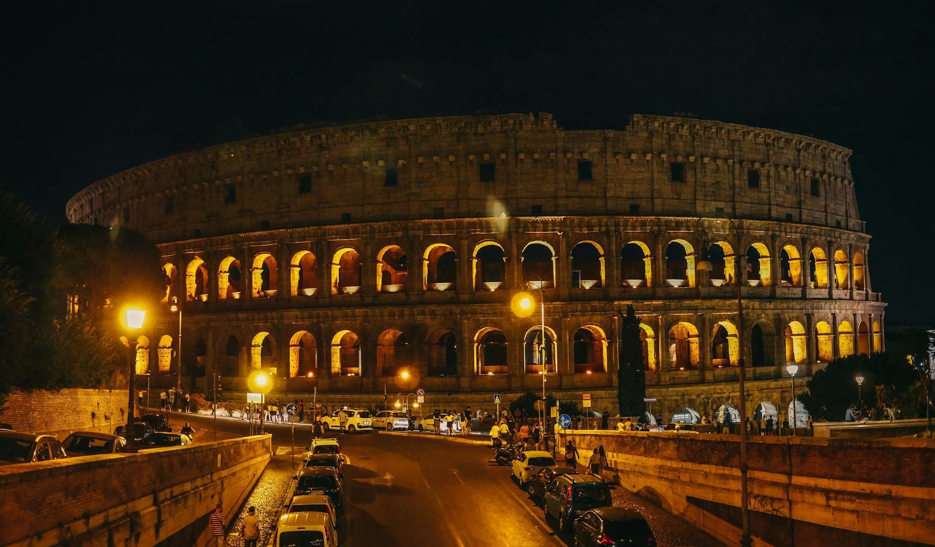 Wide view of the Colosseum in Rome illuminated at night, with glowing arches and cars parked on the street in front.