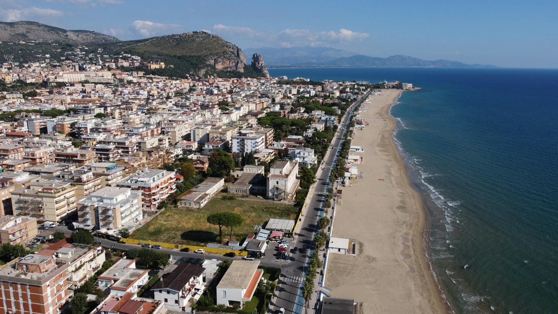 Aerial view of a picturesque Italian beach town with a long sandy shoreline, sparkling blue waters, and hills in the background.