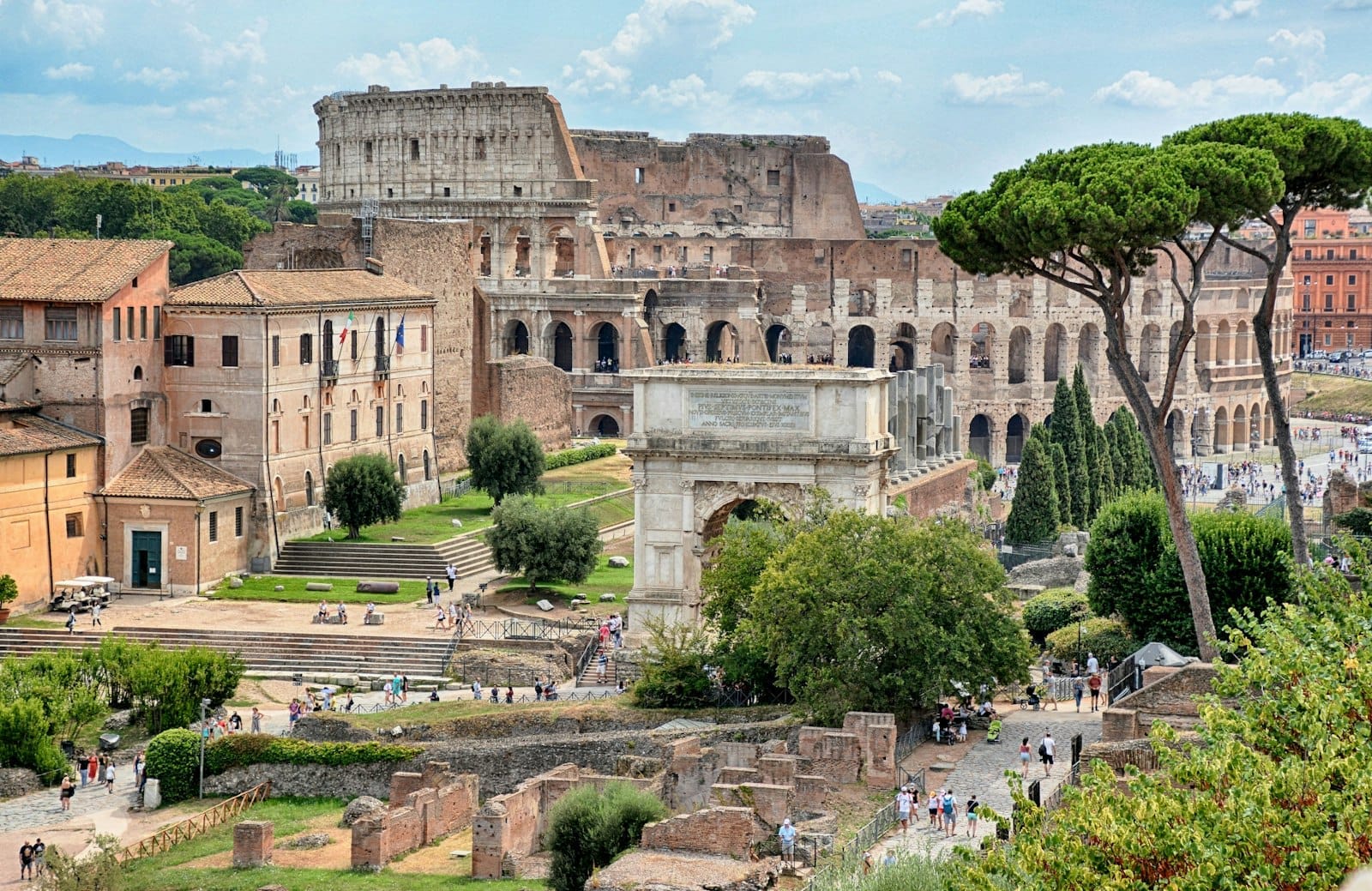 View of the Colosseum and Arch of Constantine framed by Roman ruins and umbrella pines under a bright sky, evoking Rome’s ancient heritage and day trip vibe.