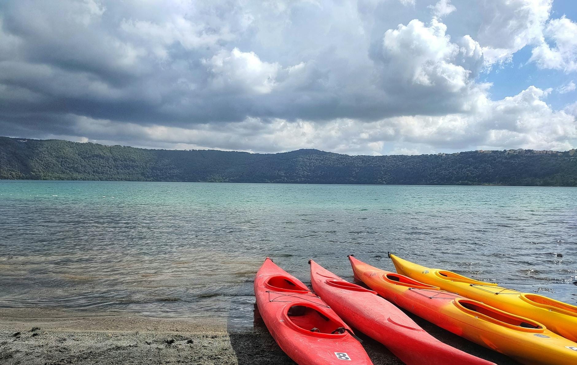 Shoreline of a volcanic lake with several colorful kayaks on the sand, forested hills in the distance and dramatic clouds overhead.