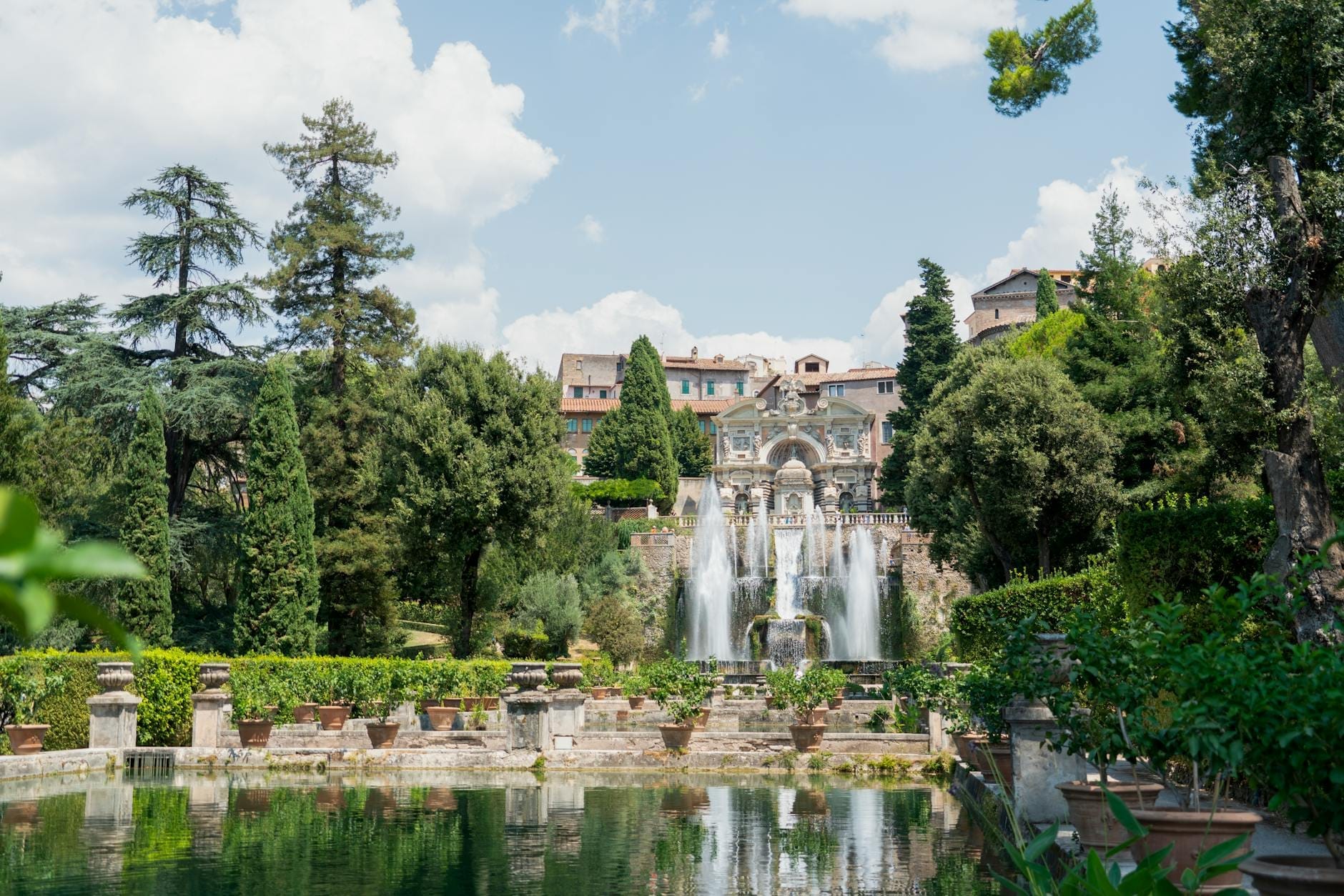 Terraced Italian Renaissance garden with large fountains, lush greenery, and a historic villa in the background under a bright sky.
