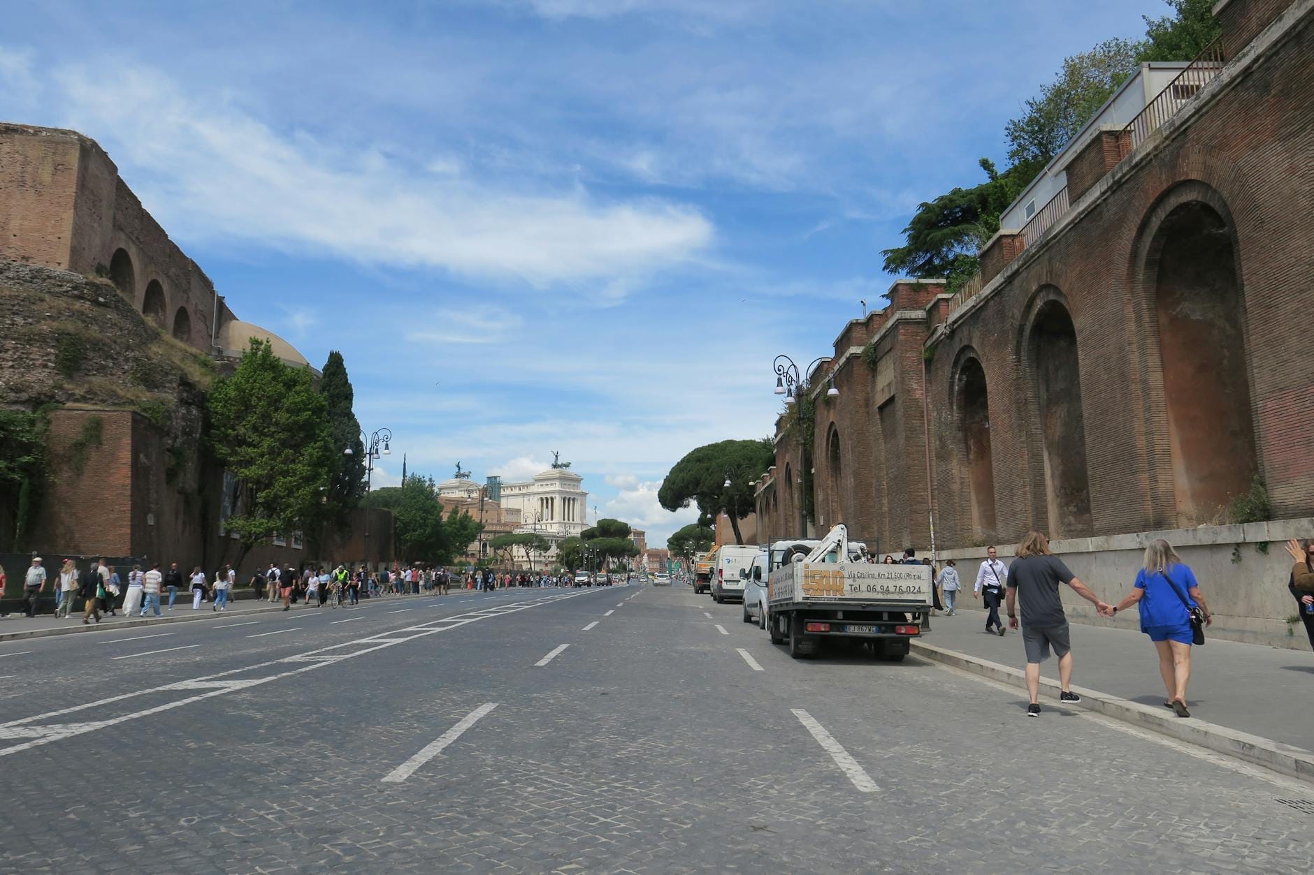 Wide cobblestone street lined with ancient Roman brick walls, distant monument, pedestrians and vehicles under blue sky in Rome.