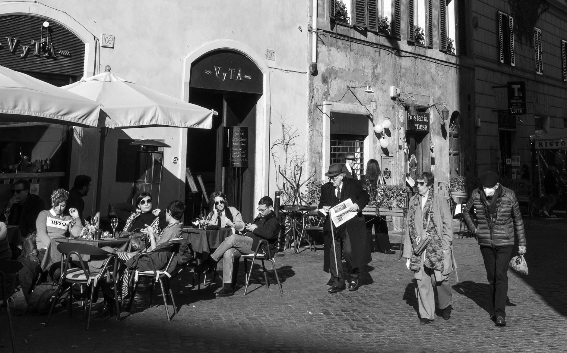 Black and white photo of people dining at outdoor tables near historic buildings, evoking the charm of Rome’s Jewish Ghetto.