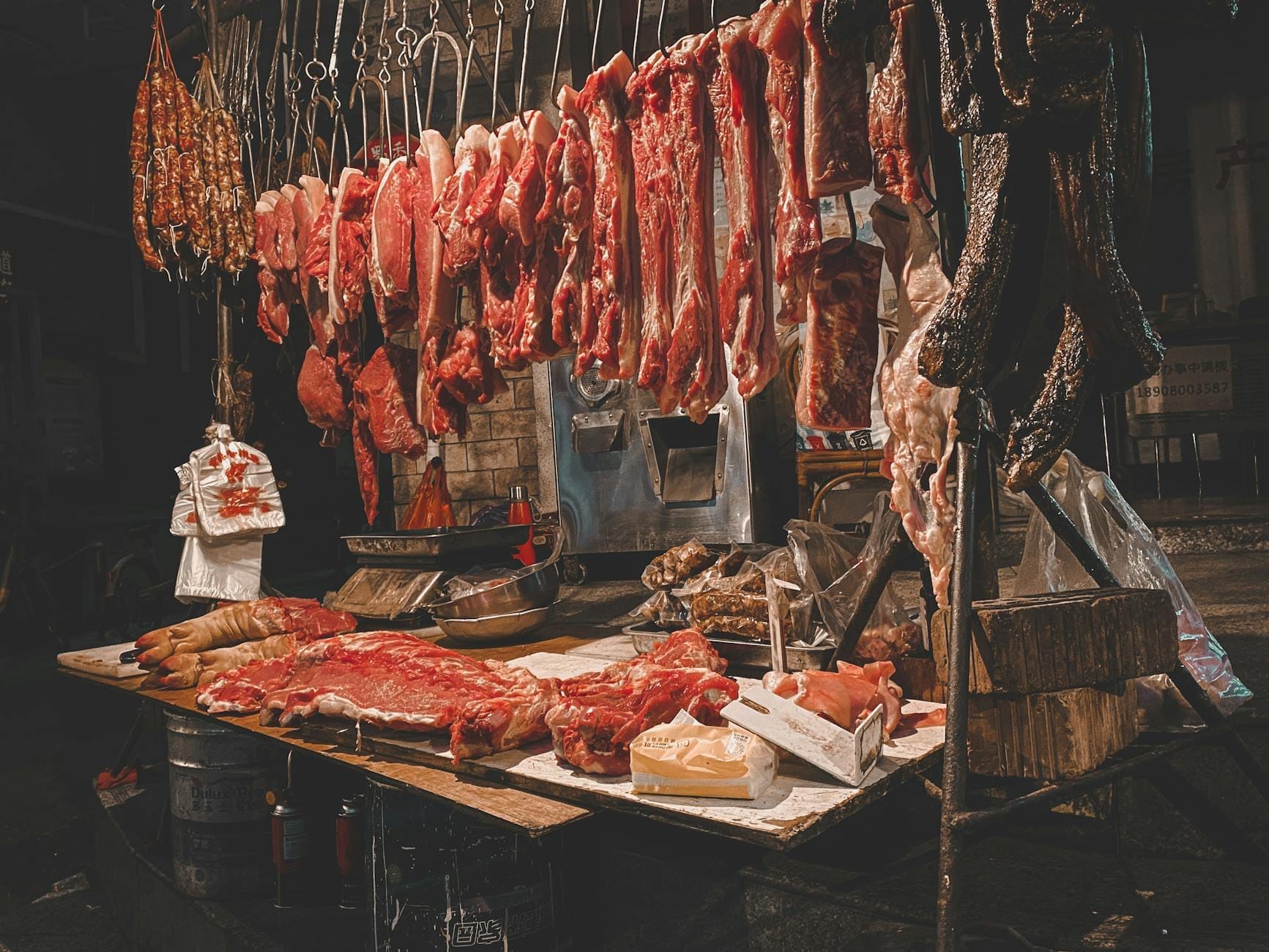 Butcher stall with various cuts of meat and offal hanging and displayed, illuminated in a market-like atmosphere.