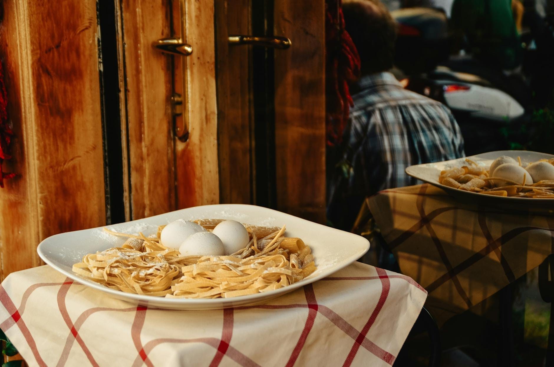 Close-up of a traditional Roman carbonara pasta dish on a checkered tablecloth outside a rustic restaurant in Rome.