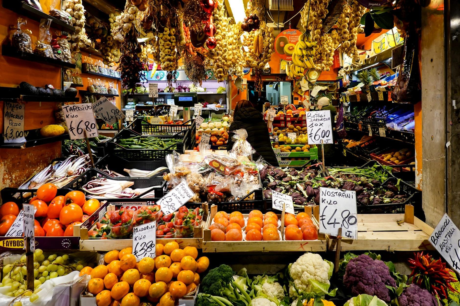 Colorful display of seasonal fruits and vegetables with price signs in a busy Roman market stall.