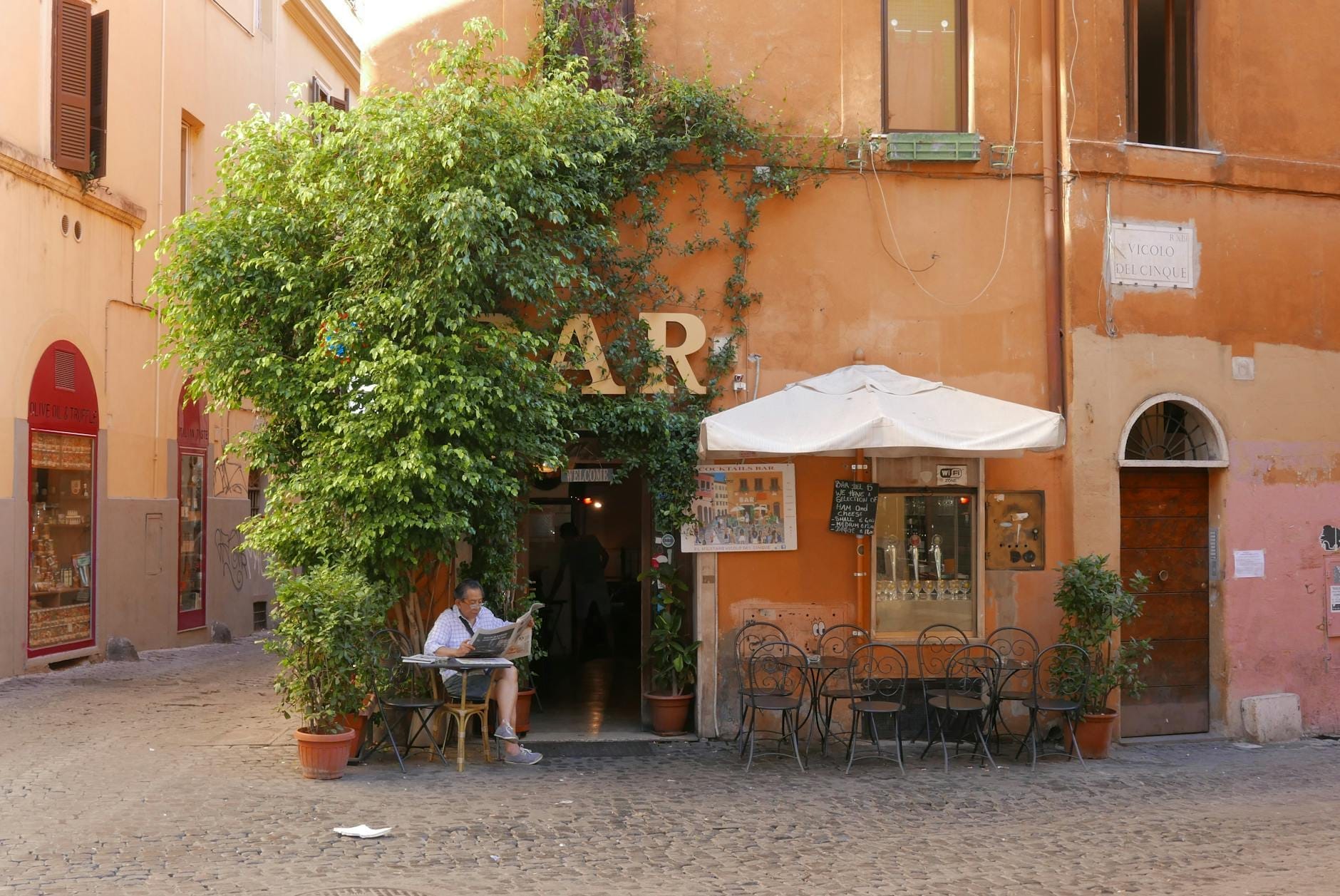 Outdoor view of a charming Roman bar with seating on a cobblestone street, orange walls, and a person enjoying coffee at a table.