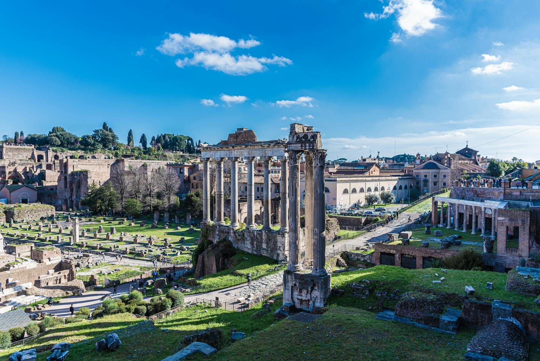 Wide view of the Roman Forum ruins in Rome under blue sky, showcasing ancient columns and remnants of historic Roman structures.