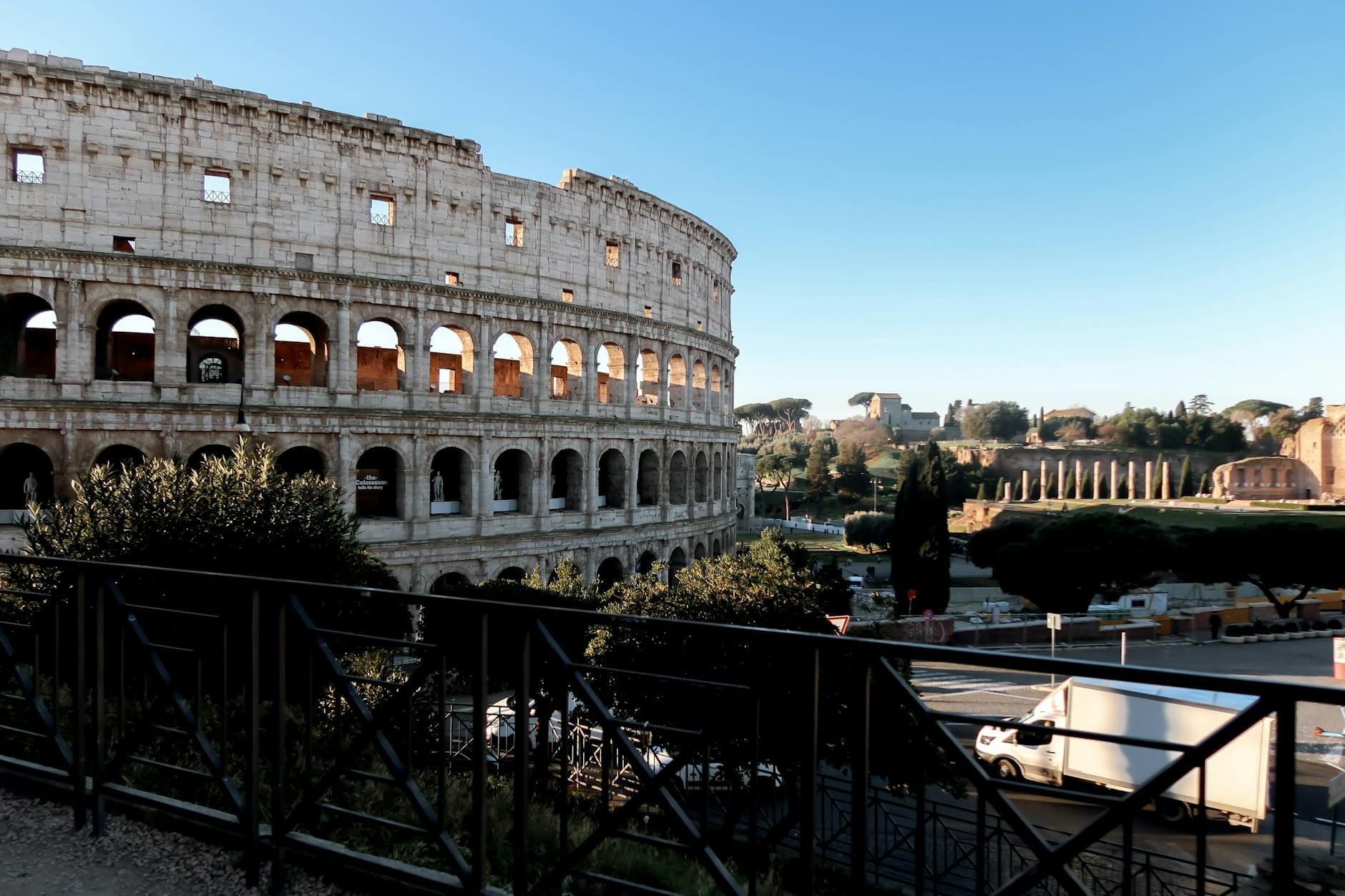 Wide daytime view of the Colosseum in Rome with the Roman Forum visible in the background under a clear blue sky.