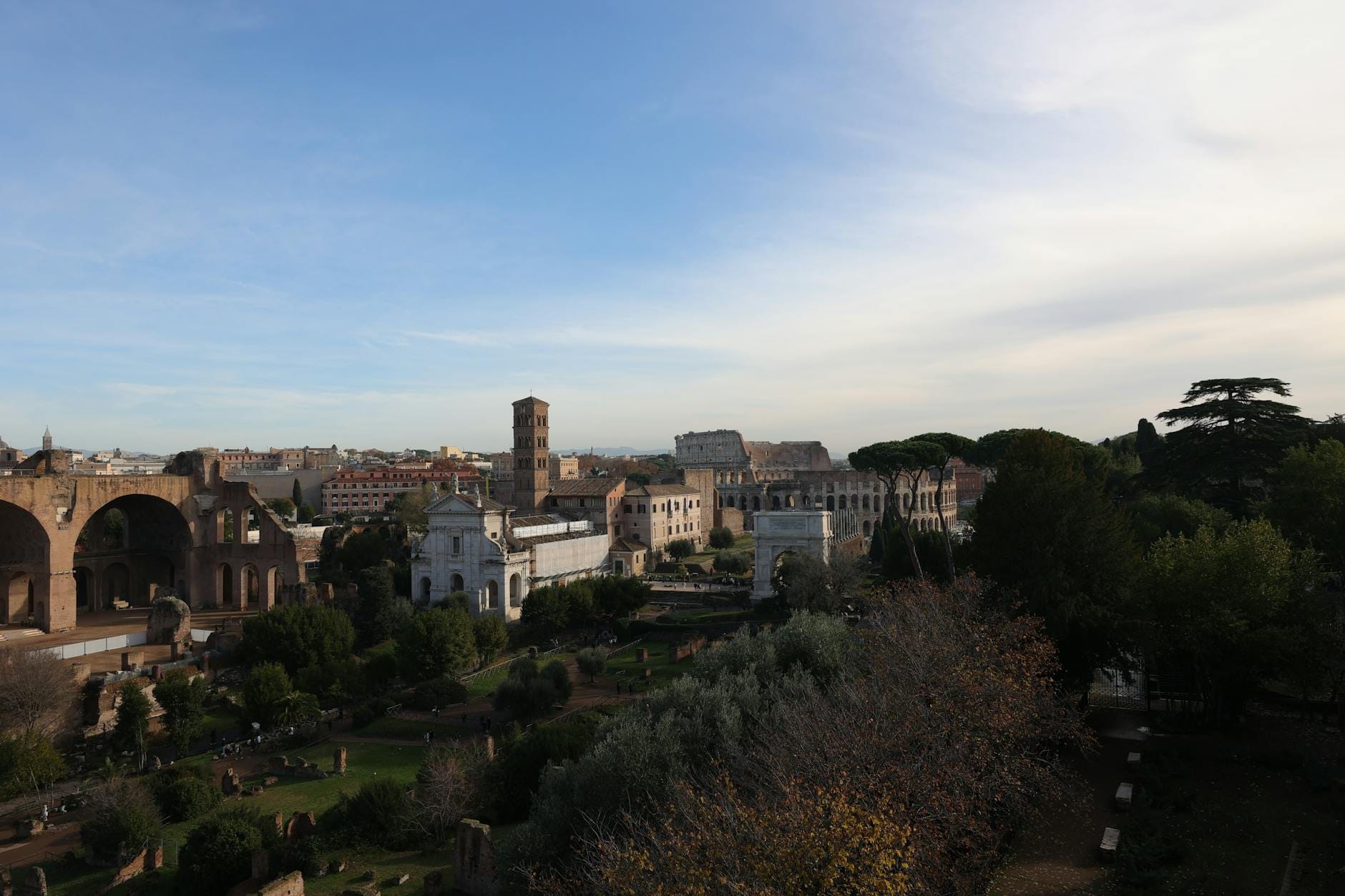 Wide view over the Roman Forum with ruins, greenery, and cityscape in the background under a blue sky.