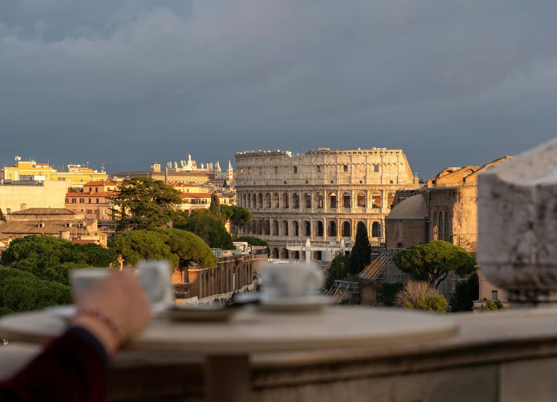 A sweeping view of Rome with the Colosseum in the background and a hand reaching for a coffee cup in the foreground on a sunny day.