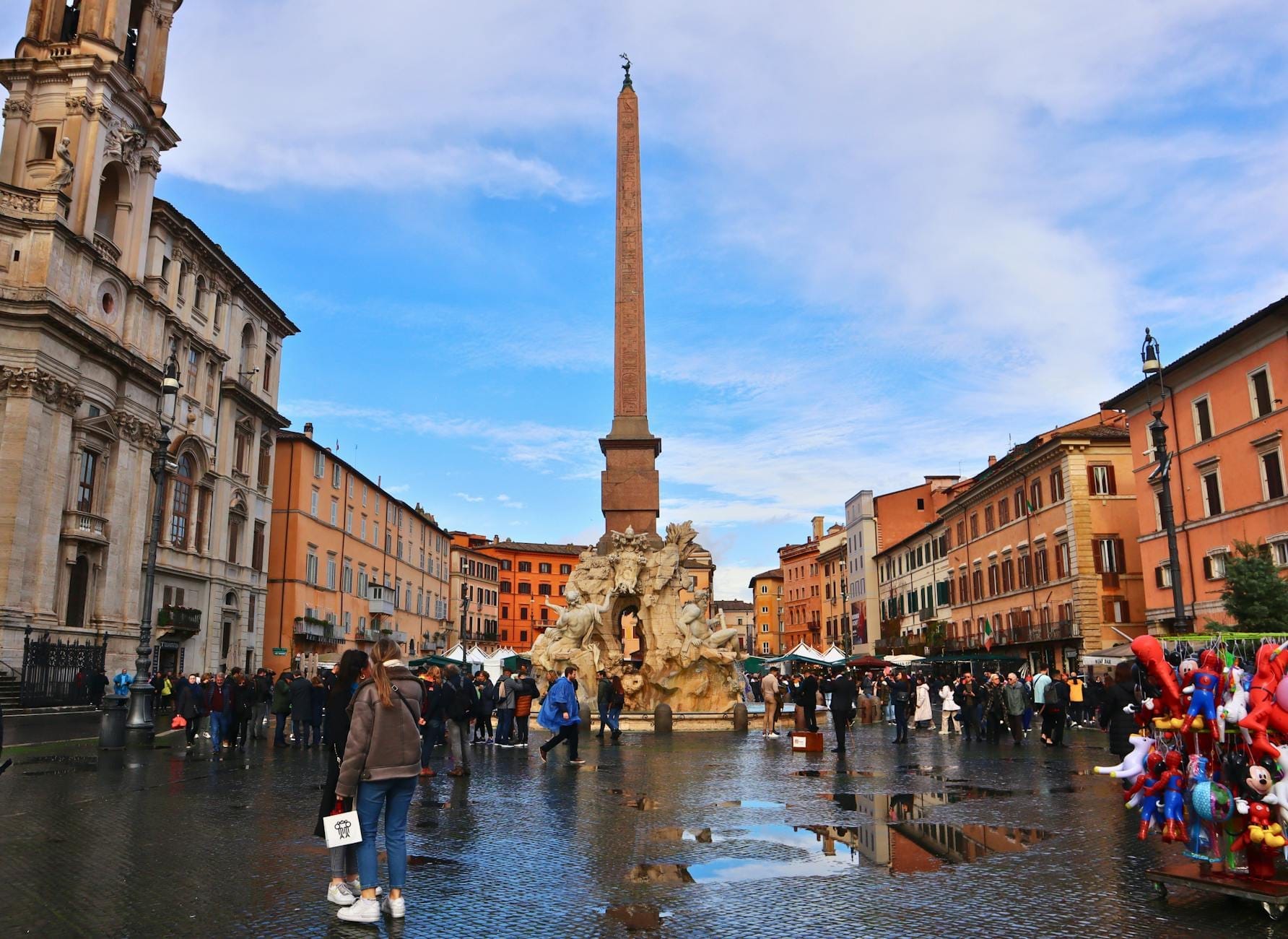 Wide view of Piazza Navona in Rome with crowds, fountains, central obelisk, colorful buildings, and baroque church under a bright blue sky.