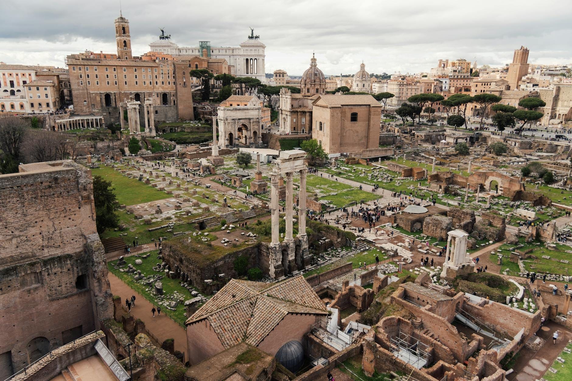 Wide view of the Roman Forum archaeological site, with ancient ruins and landmark buildings under a cloudy sky in Rome, Italy.