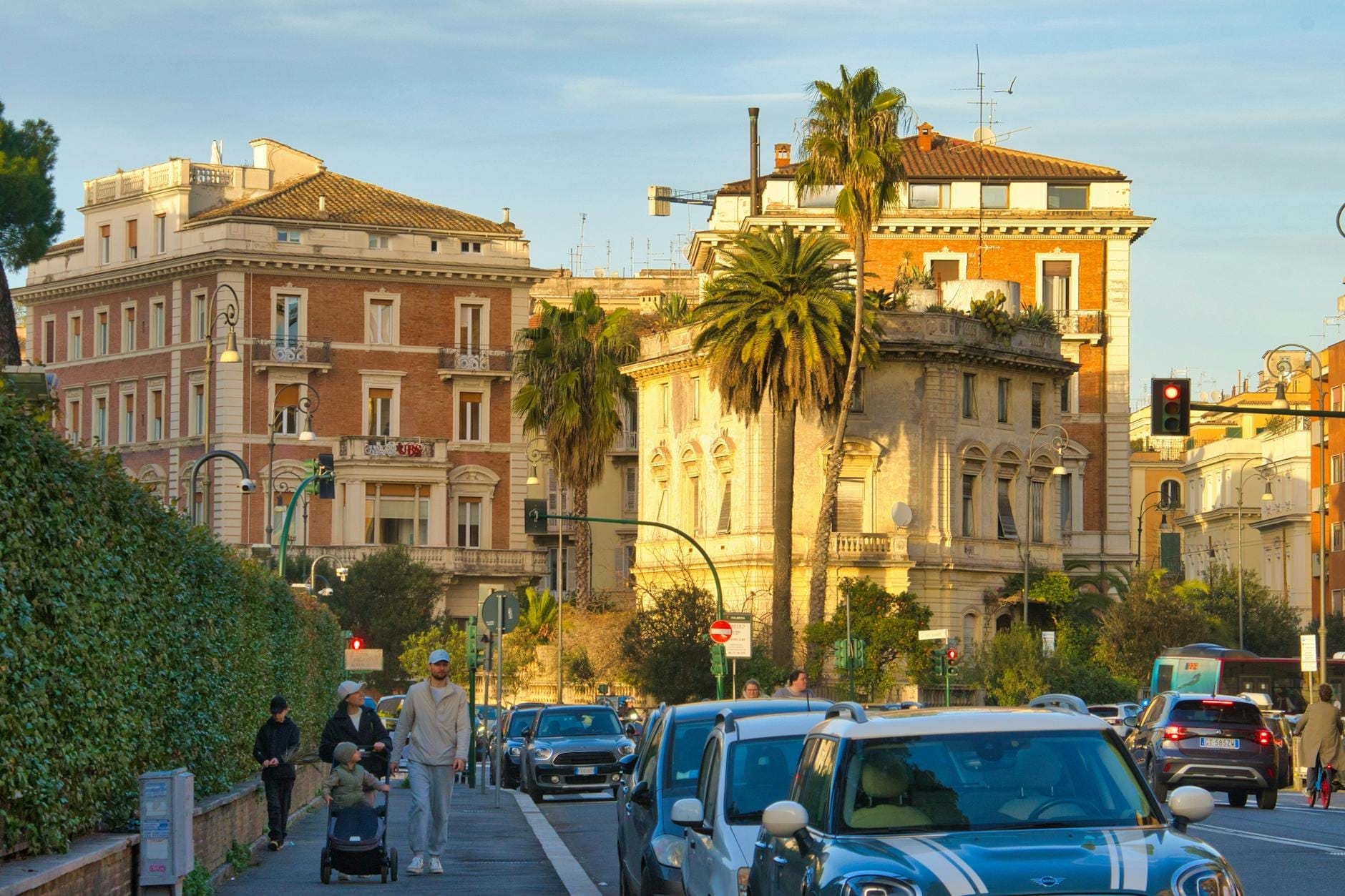 Street in Rome with cars, palm trees, historic buildings, and people including a parent pushing a stroller on the sidewalk.
